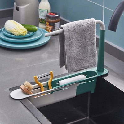 Adjustable telescopic sink rack in green and white, mounted over a kitchen sink, holding a towel, sponge, brush, and utensils for organized storage.